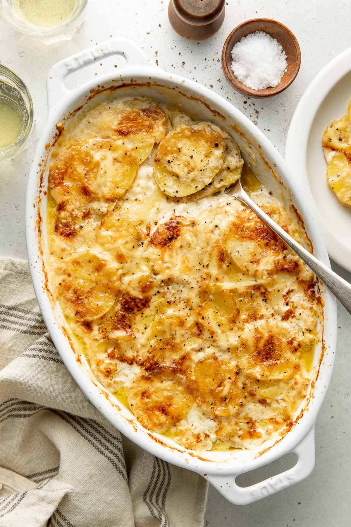 An oval baking dish of golden, cheesy scalloped potatoes, with a spoon scooping some out. A plate with a serving, salt, pepper, and striped napkin are nearby on a light surface.