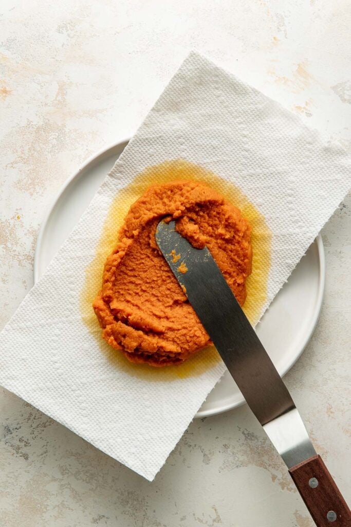 A metal spatula spreads orange miso paste on a white paper towel laid over a white plate, with oil soaking into the towel.