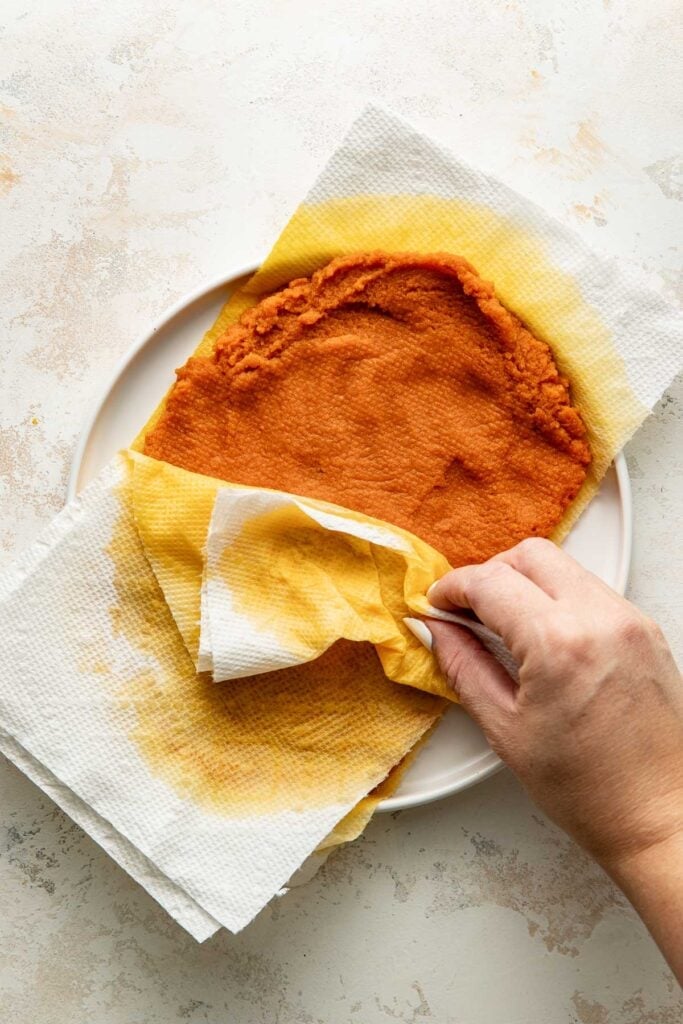 A hand lifts a paper towel off a round, orange food item on a plate, with the towel showing oil stains.