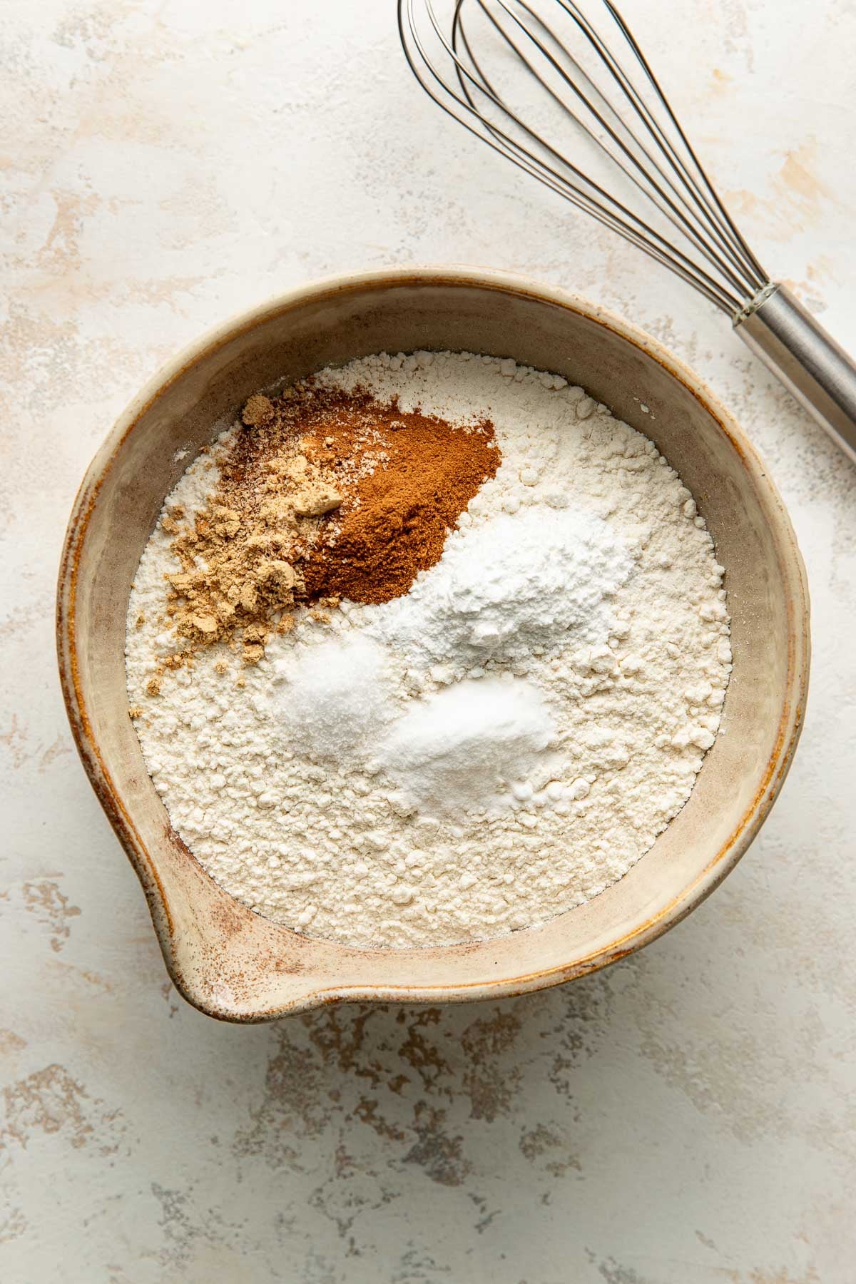 A bowl filled with flour, ground ginger, cinnamon, baking soda, and baking powder sits on a light surface next to a metal whisk.