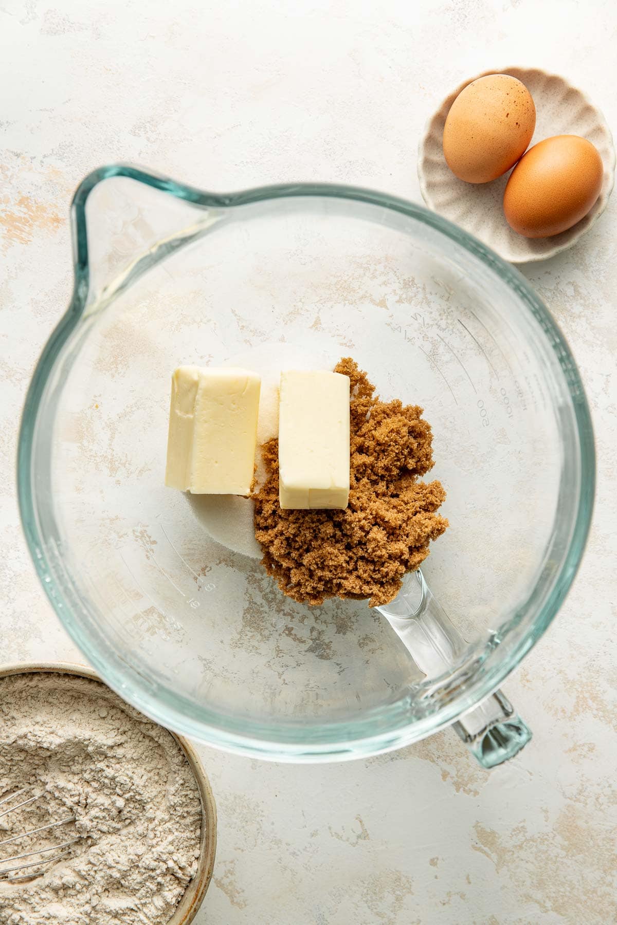 A glass mixing bowl with two sticks of butter and brown sugar. Nearby are a small bowl of flour and a plate with two eggs, all on a light textured surface.