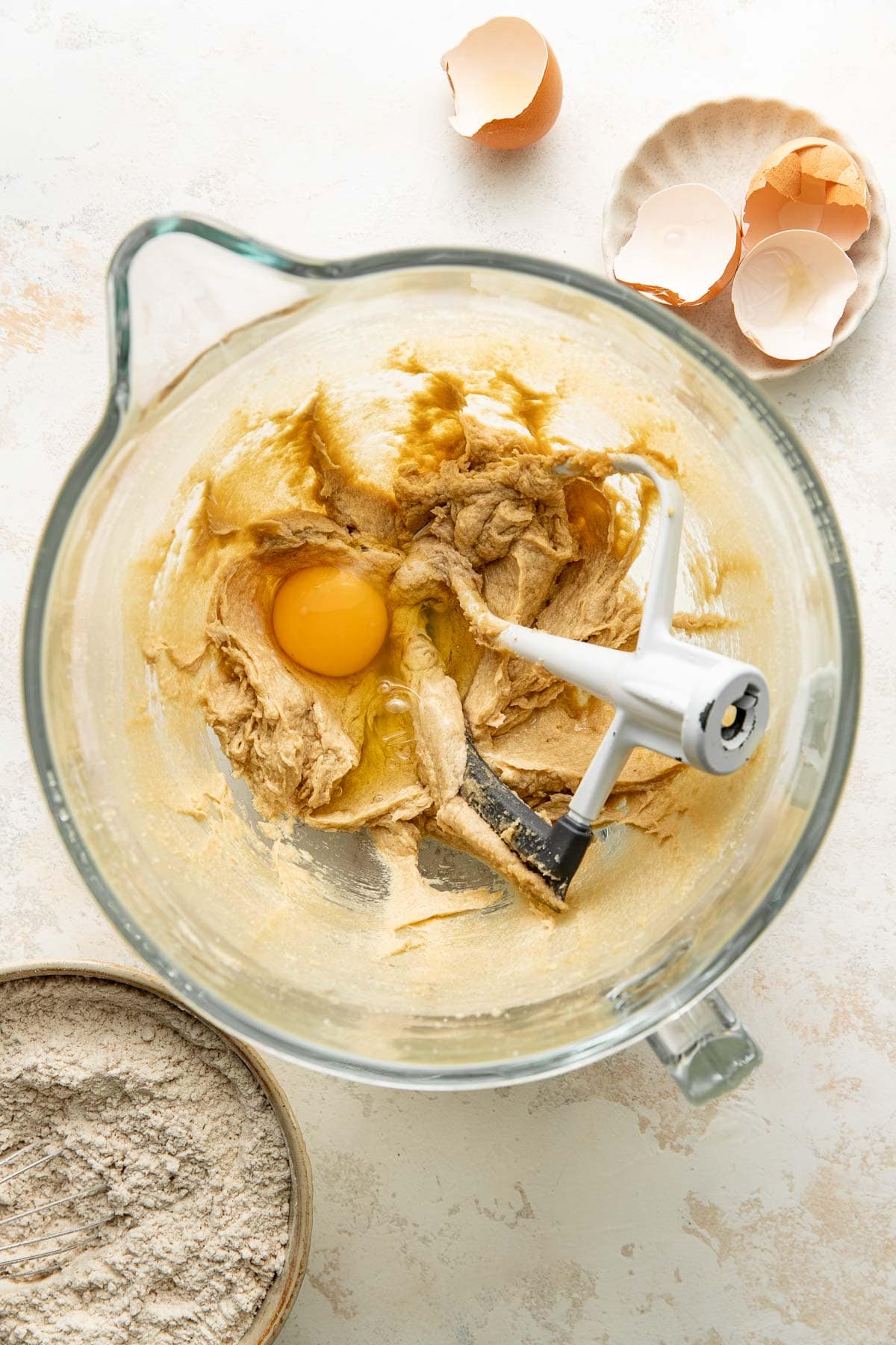 A mixing bowl with cookie dough, an egg in the center, and a stand mixer paddle. Nearby are a bowl of flour and an egg shell in a small dish on a light countertop.