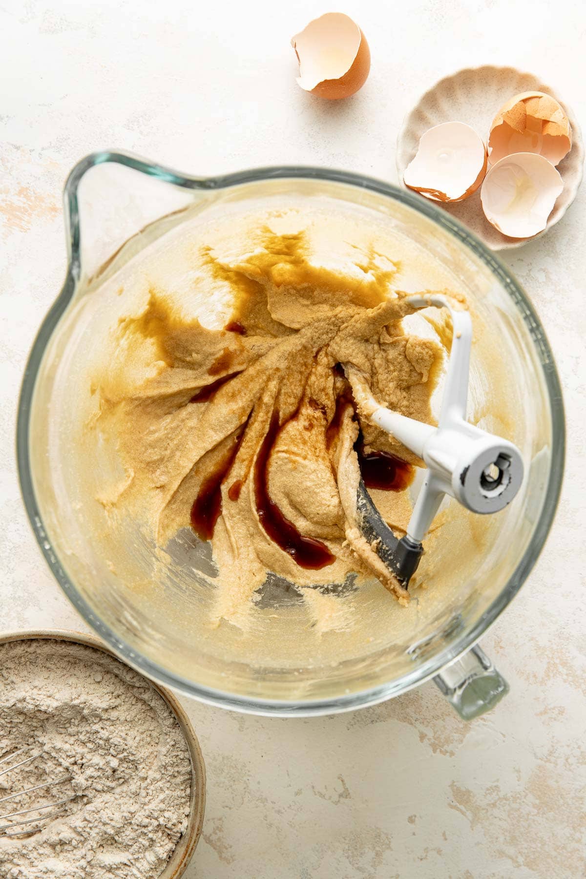 Glass mixing bowl with cookie dough, vanilla extract, and a stand mixer paddle; nearby are cracked eggshells in a dish and a bowl of flour.