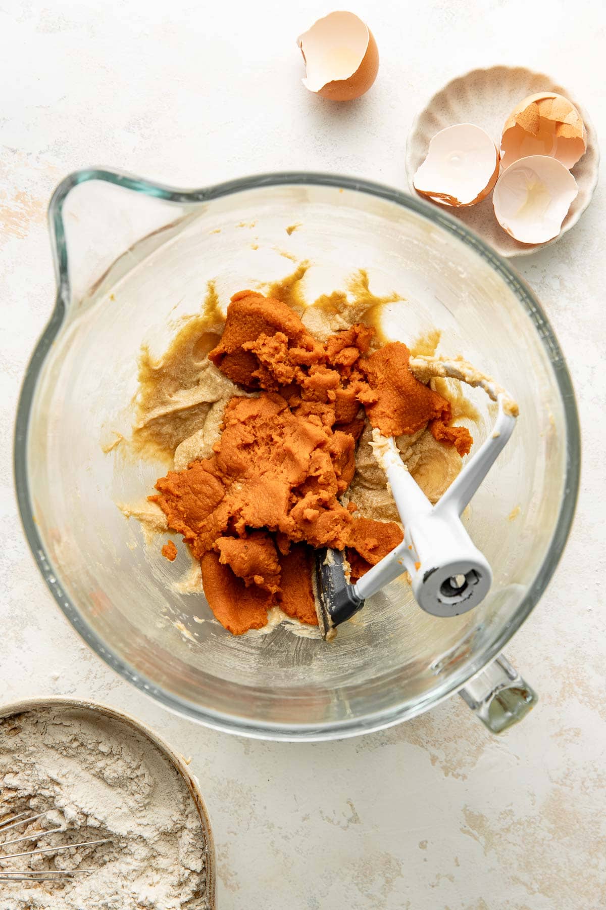 A glass mixing bowl with brown cookie dough and a paddle attachment, next to a bowl of eggshells and a plate of flour on a white countertop.
