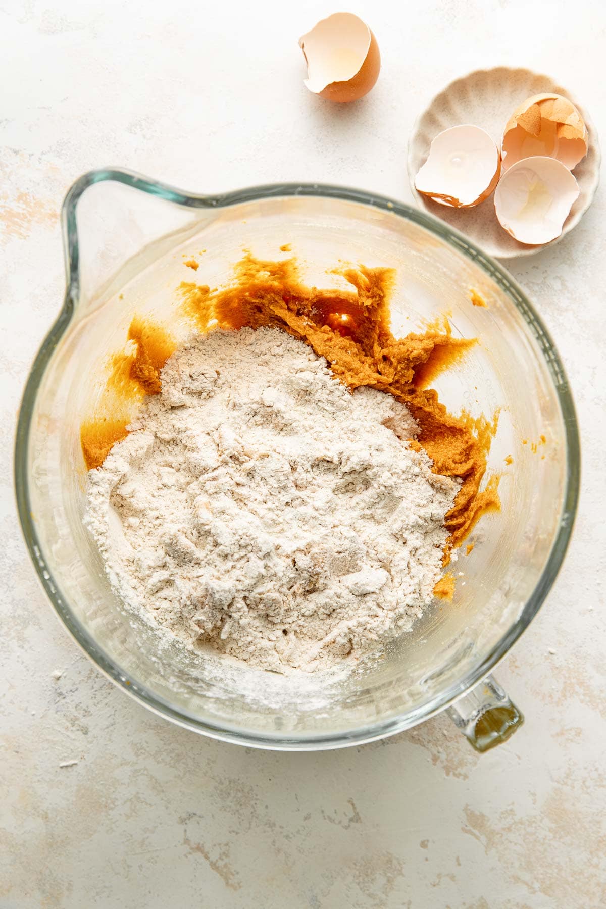 A glass mixing bowl with brown sugar and flour mixture sits on a light surface. Nearby, a small dish holds cracked eggshells, and another eggshell rests on the counter.