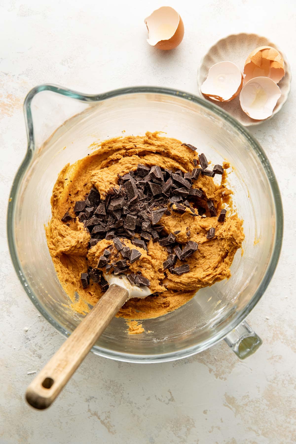 A glass bowl with cookie dough and chocolate chunks being mixed with a spatula. Broken eggshells are in a small dish nearby on a light surface.