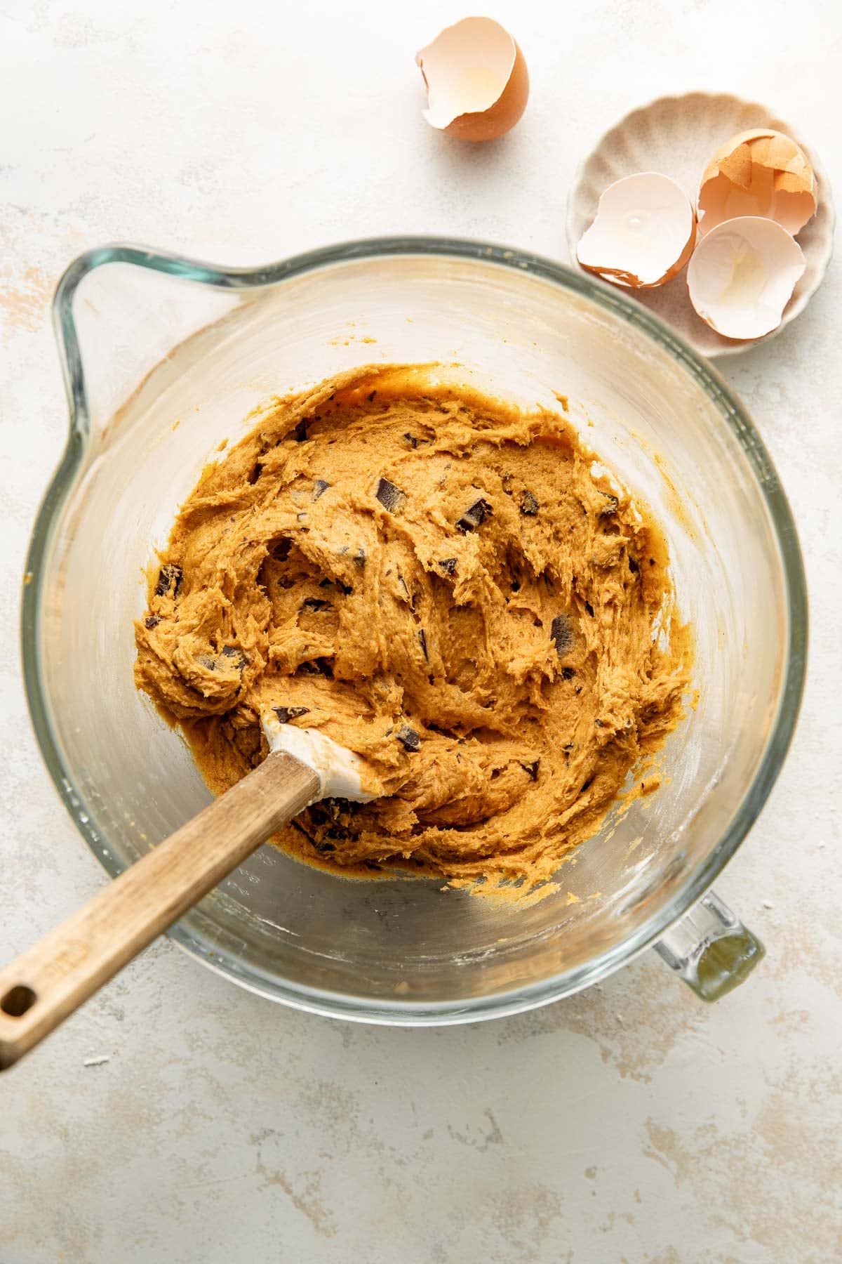 A glass mixing bowl with chocolate chip cookie dough and a wooden spatula, next to a small dish holding cracked eggshells on a light countertop.