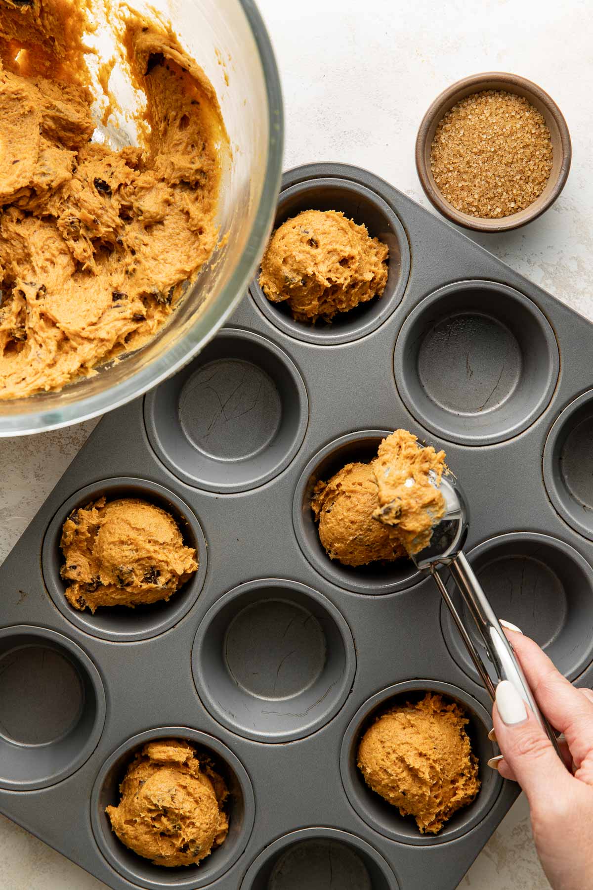 A hand uses a scoop to place cookie dough into a muffin tin, with a glass bowl of dough and a small bowl of brown sugar beside it.