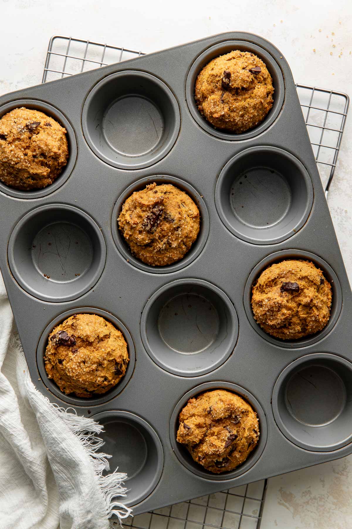 A muffin tin with seven chocolate chip muffins, placed on a wire cooling rack with a white cloth nearby. Some muffin cups are empty.