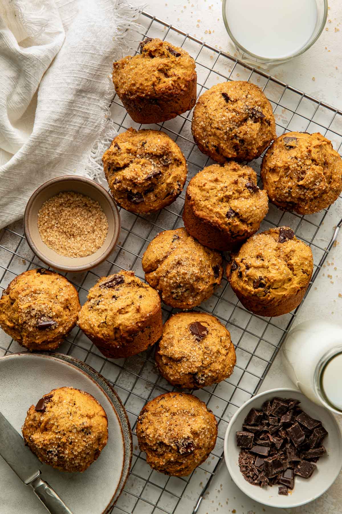 Twelve chocolate chip muffins on a cooling rack, with bowls of sugar and chocolate chunks, two glasses of milk, and a plate with a knife nearby.