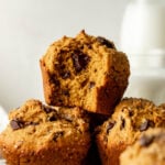 A stack of chocolate chip muffins, one with a bite taken out, sits in front of a blurred glass bottle of milk on a white surface.