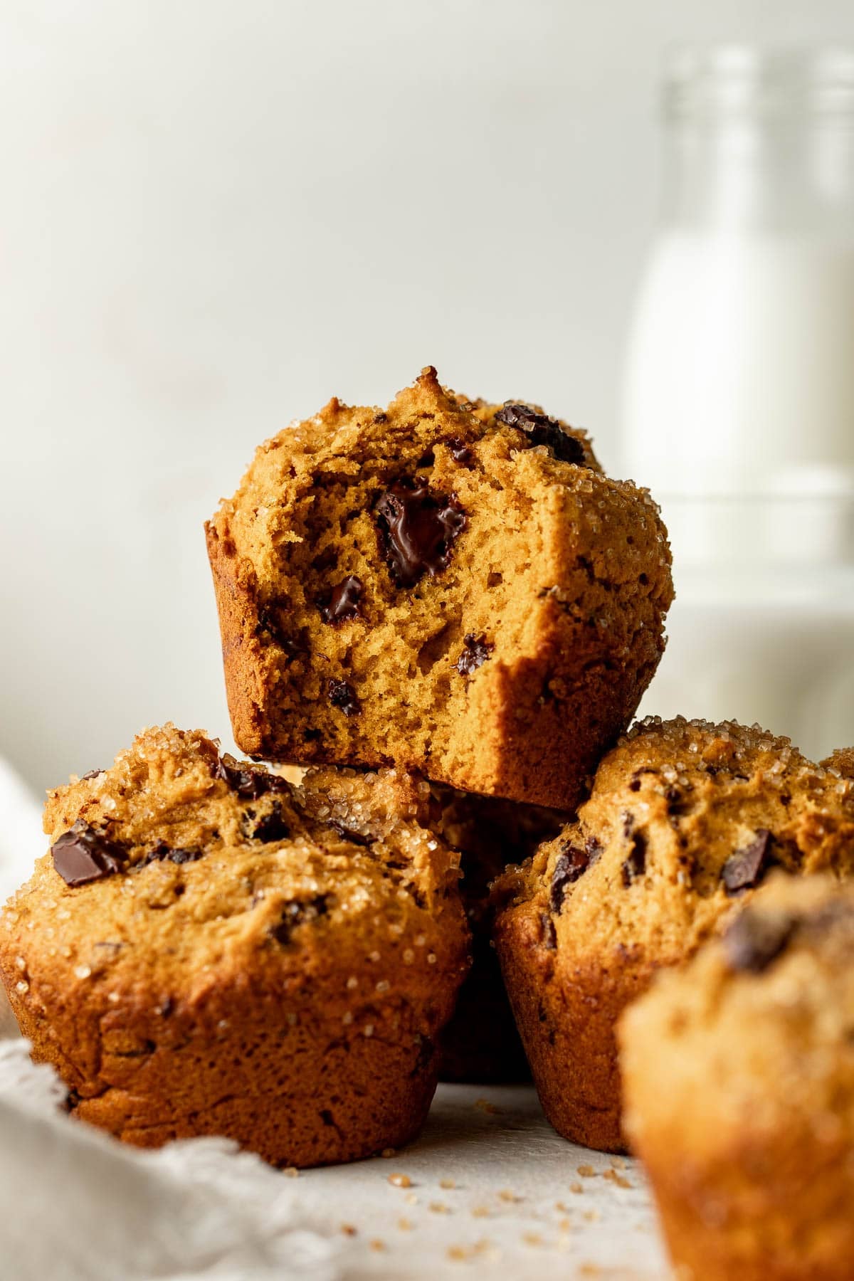 A stack of chocolate chip muffins, one with a bite taken out, sits in front of a blurred glass bottle of milk on a white surface.