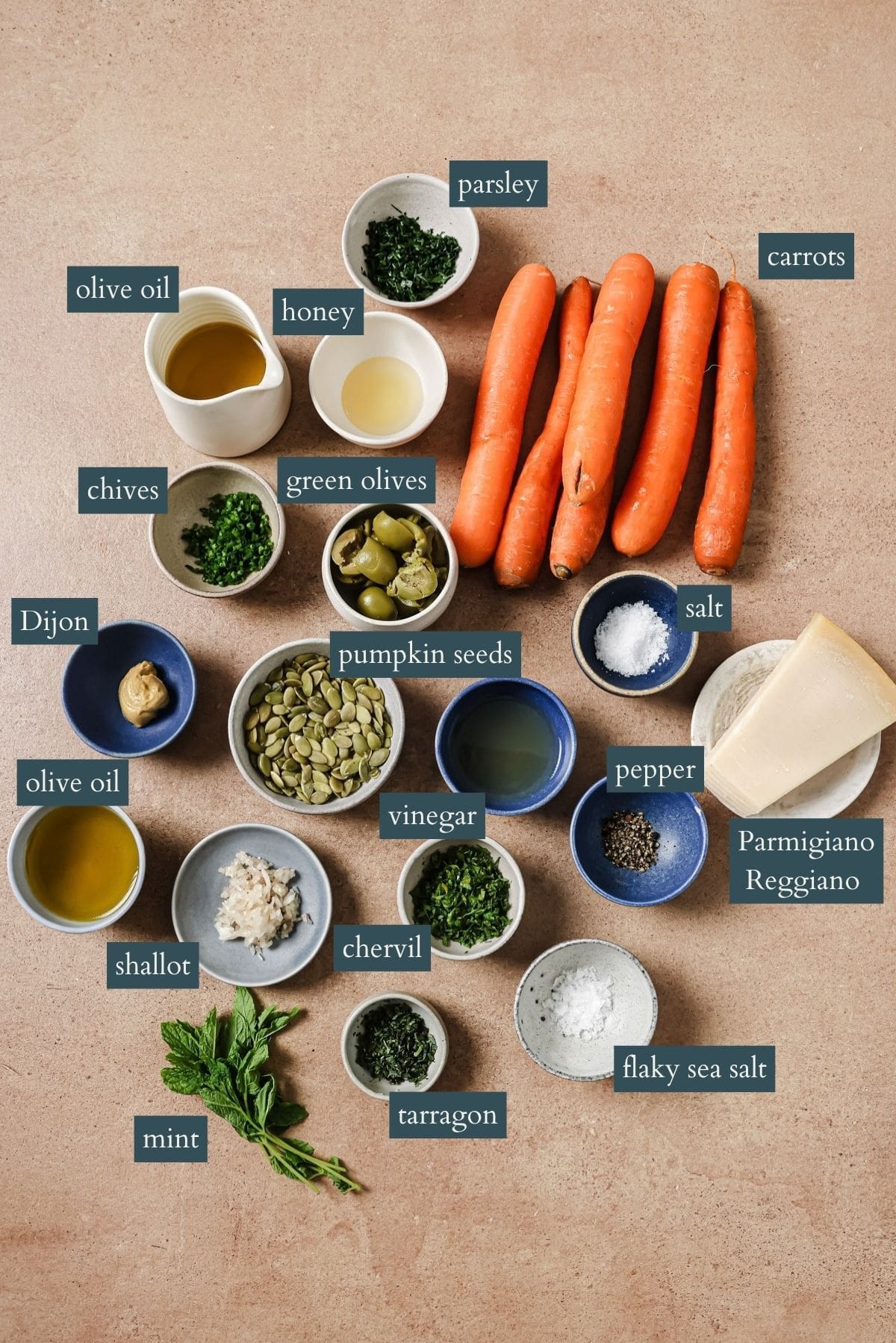 Ingredients for a carrot salad are arranged on a tabletop, including carrots, herbs, seeds, cheese, seasonings, Dijon, shallot, honey, green olives, olive oil, and bowls of salt, pepper, vinegar, and flaky sea salt. Labels identify each item.