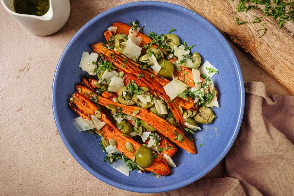 A blue bowl filled with roasted carrot slices, green olives, shaved cheese, and chopped herbs, placed on a beige surface next to a tan napkin and a wooden cutting board with more herbs.