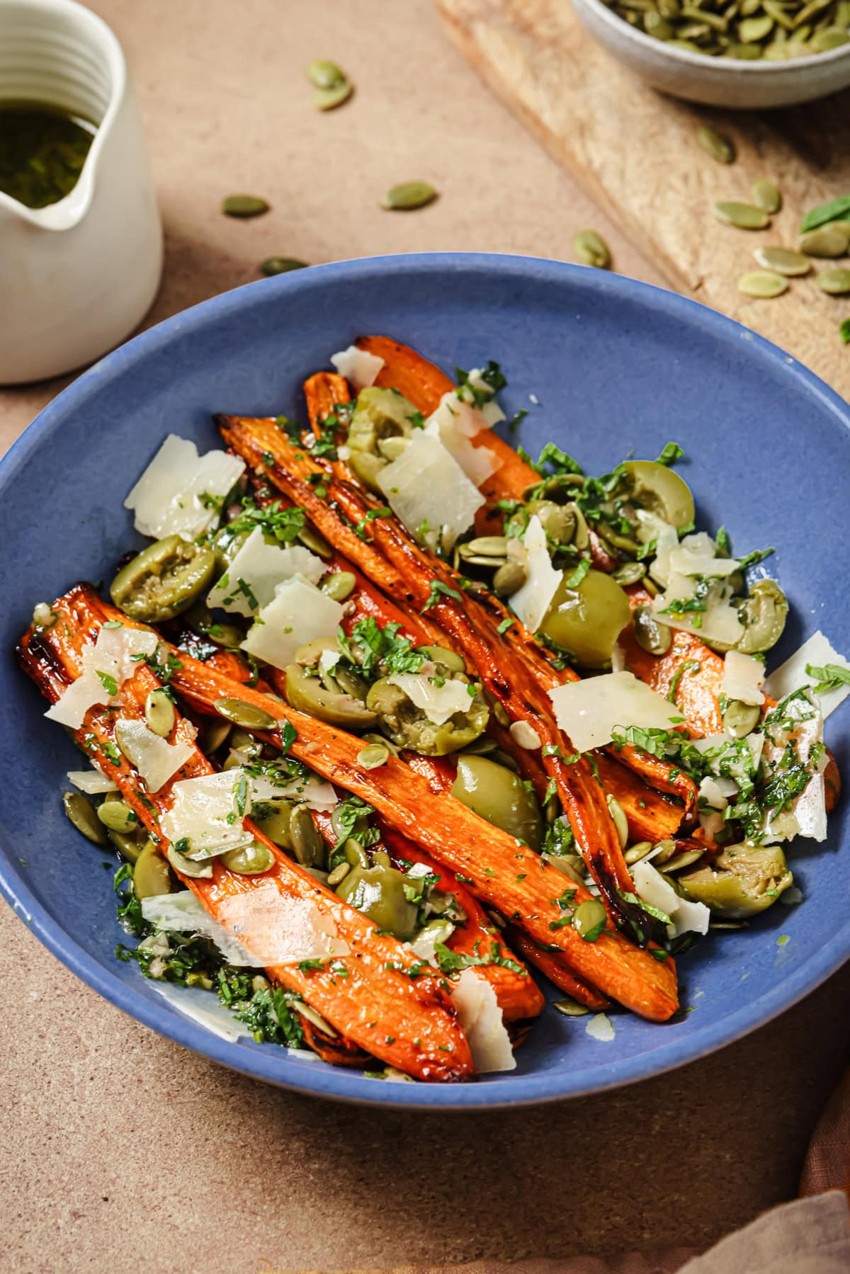 A blue bowl filled with roasted carrots, shaved parmesan, green olives, chopped herbs, and pumpkin seeds, with a white sauce pitcher and wooden board in the background.