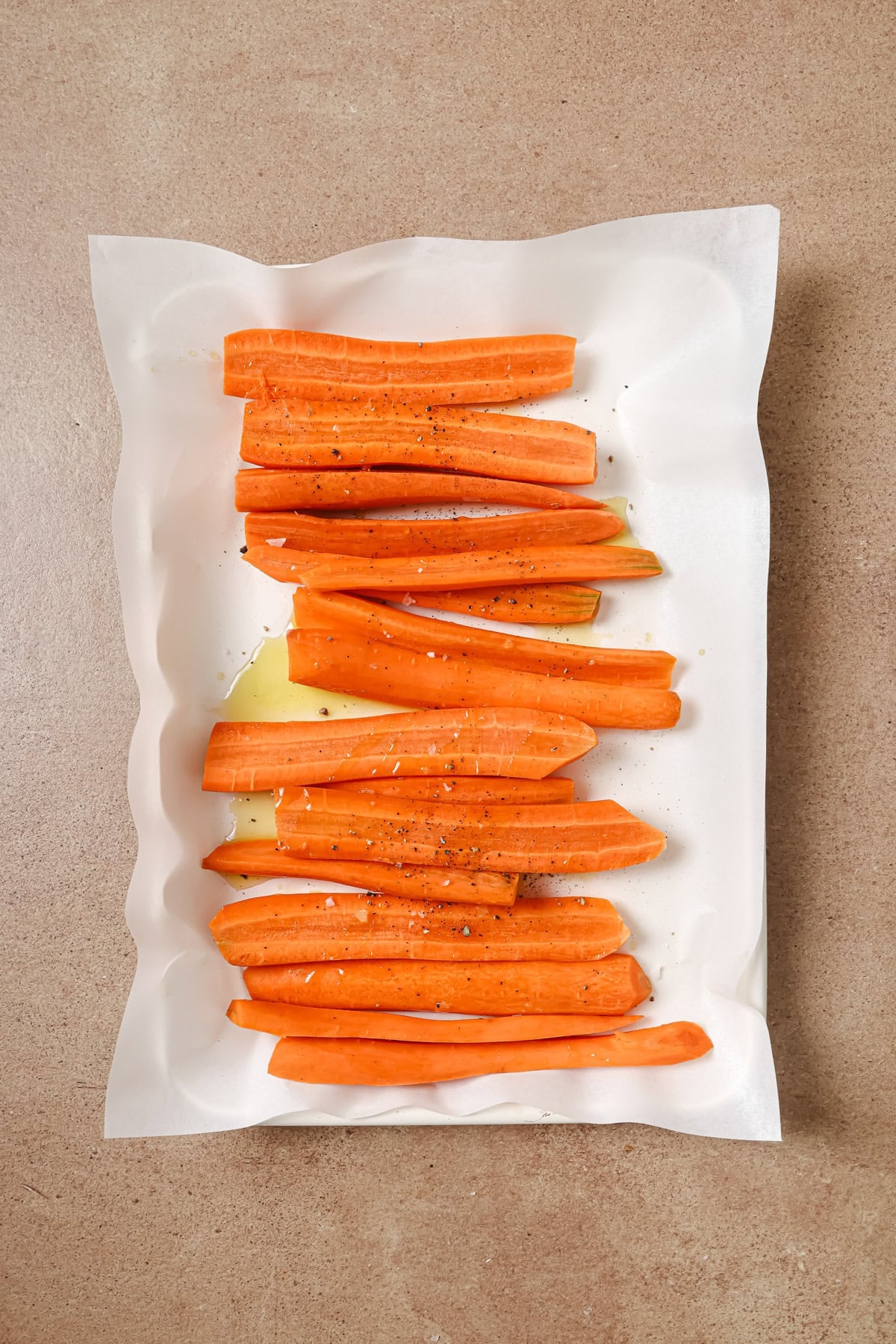 Carrot sticks on parchment paper, seasoned with salt, pepper, and olive oil, arranged neatly on a light brown countertop.