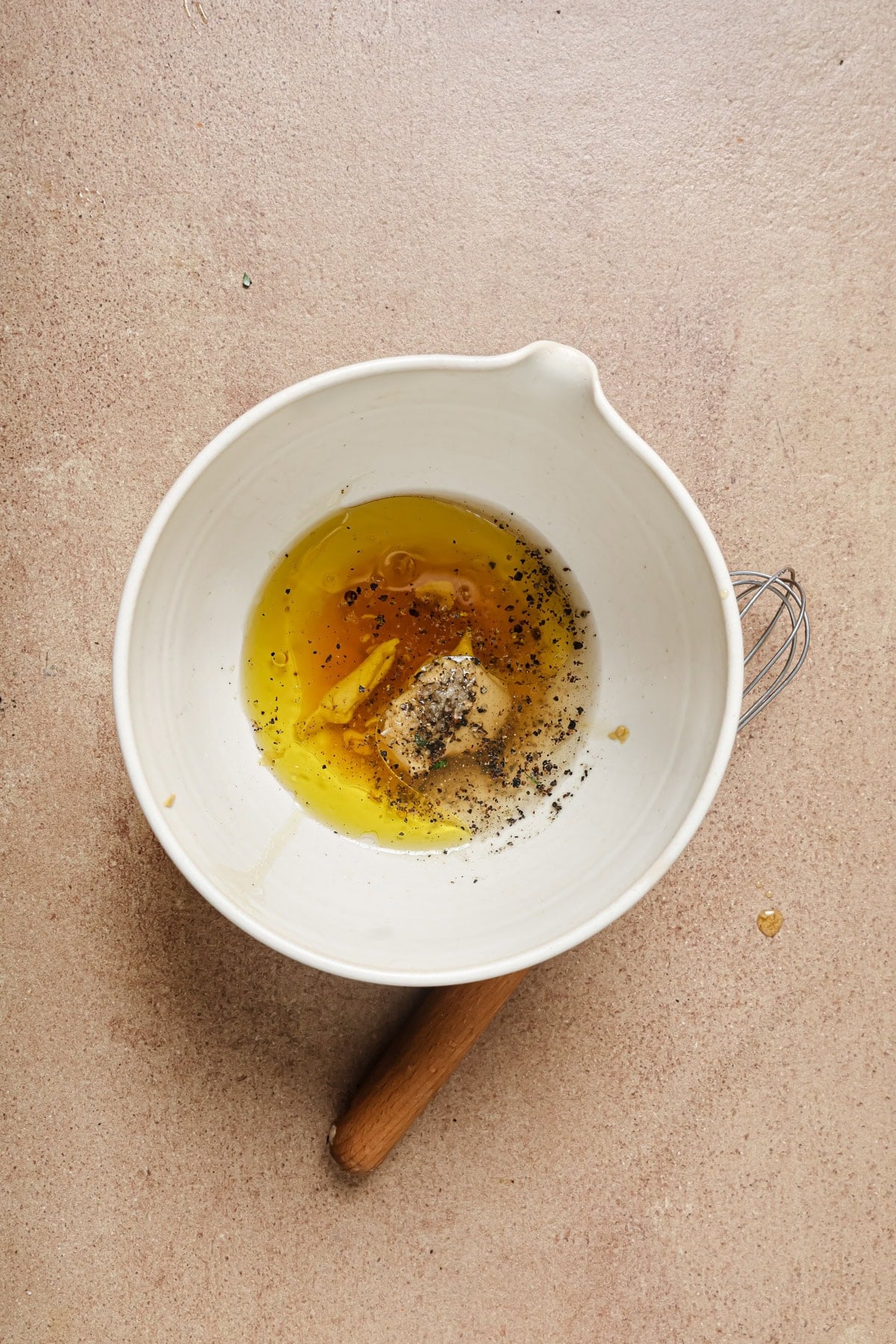 A white mixing bowl with oil, mustard, black pepper, and seasonings inside sits on a beige surface next to a whisk with a wooden handle.