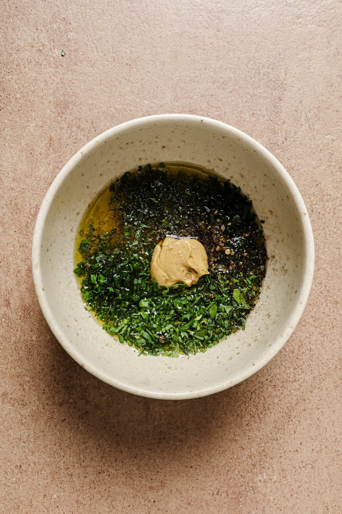 A white bowl contains chopped herbs, olive oil, a dollop of mustard, and ground pepper on a light brown surface.