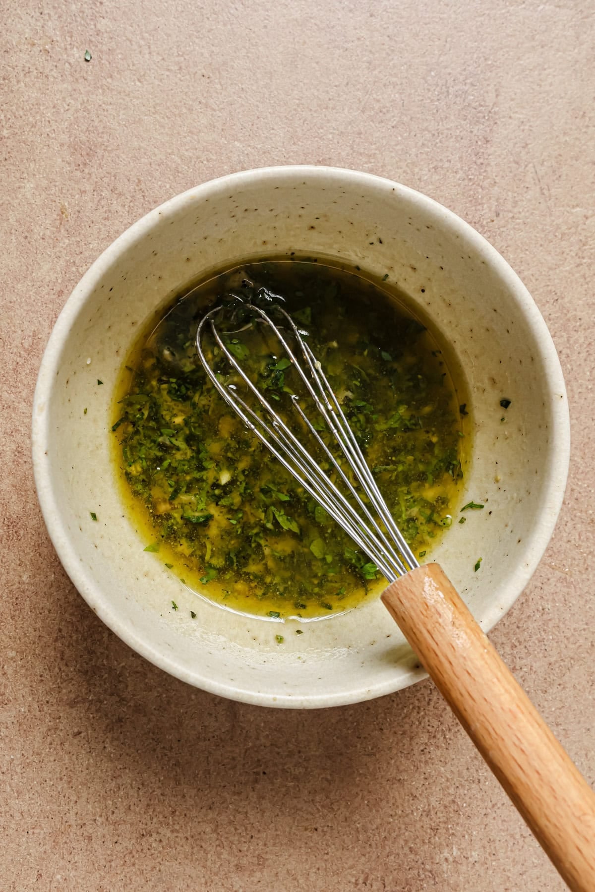 A ceramic bowl filled with a green herb dressing or marinade being mixed with a metal whisk with a wooden handle, on a light brown surface.