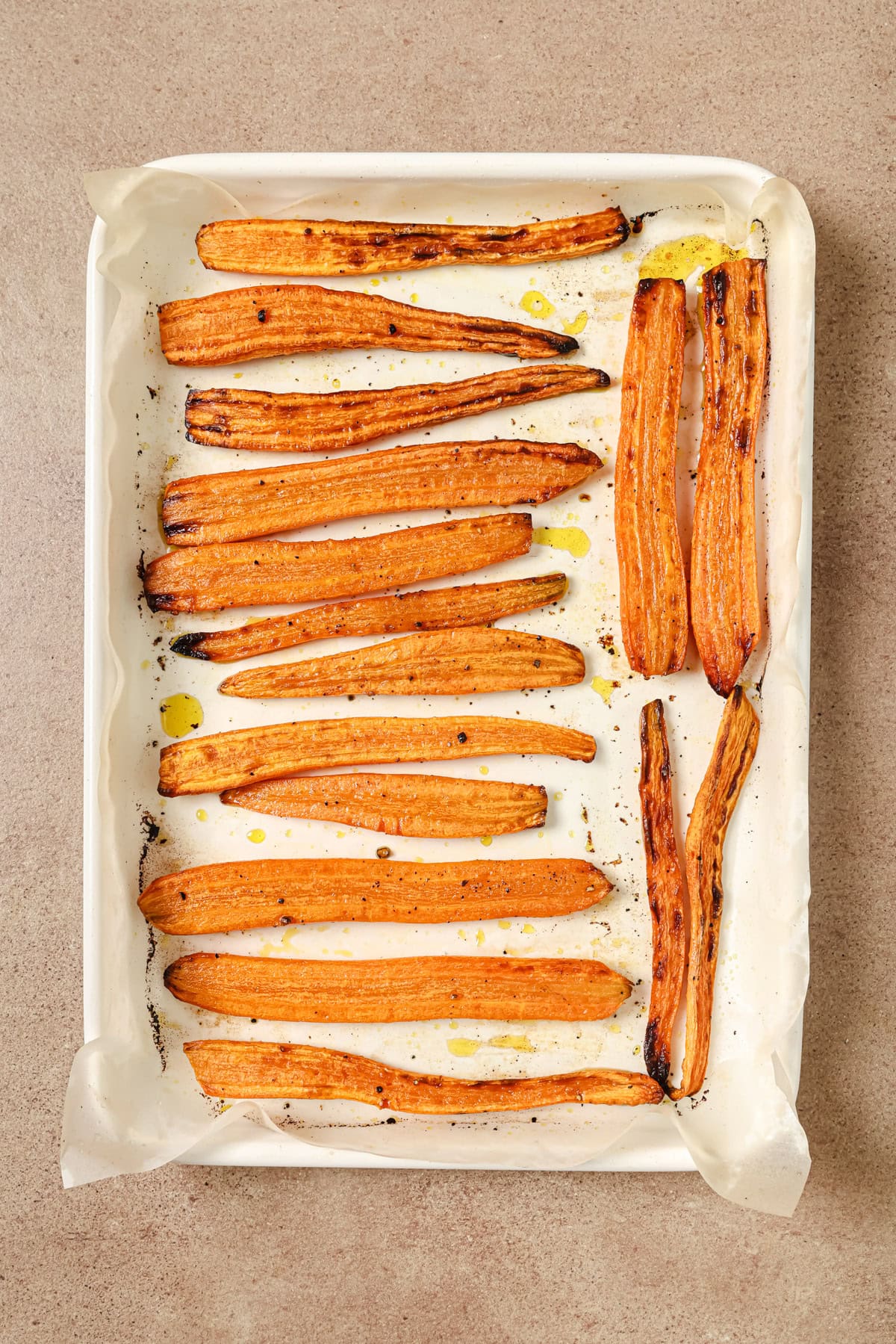 Roasted carrot halves arranged in rows on a parchment-lined baking sheet, lightly drizzled with oil and sprinkled with pepper, on a neutral countertop.