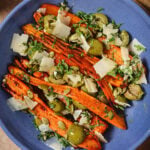 Blue bowl filled with roasted carrots, green olives, shaved cheese, seeds, and fresh herbs on a beige surface.