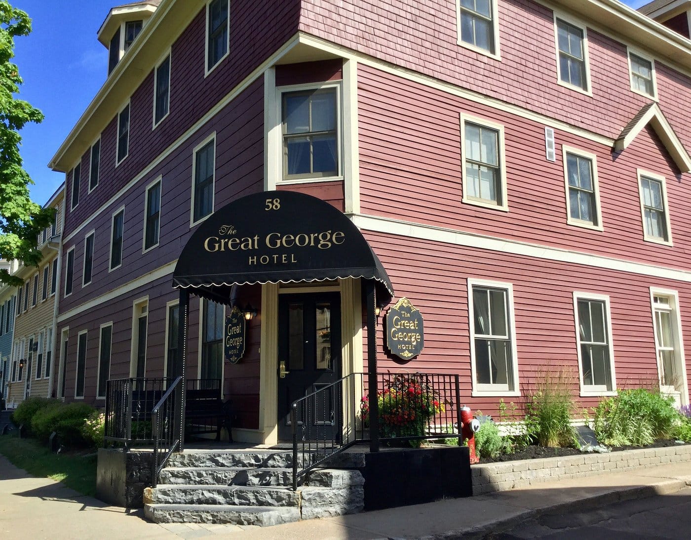 A maroon, three-story building with white trim houses The Great George Hotel. A black awning with gold lettering marks the entrance, reached by stone steps. Flowers and greenery decorate the exterior.