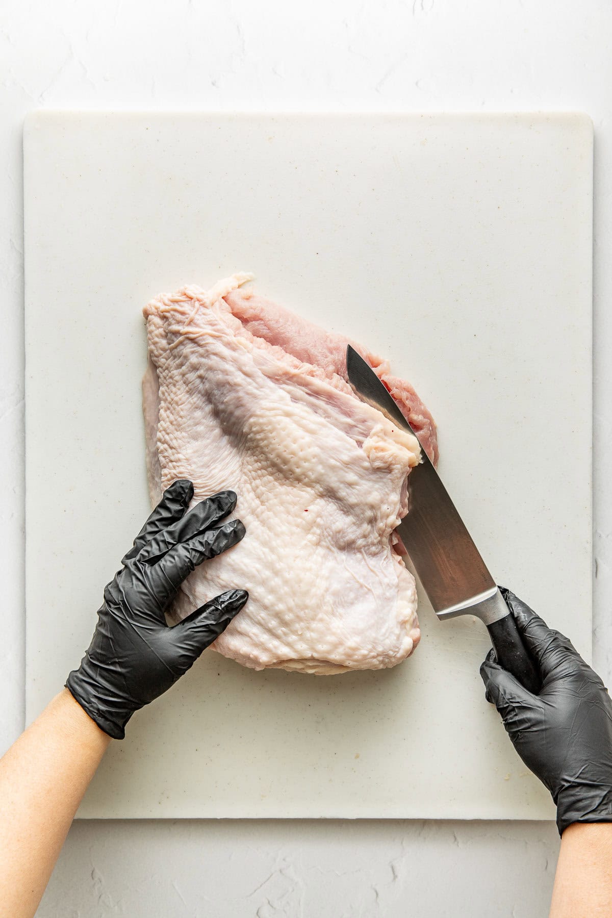 A person wearing black gloves holds a raw turkey breast on a white cutting board and slices it with a large chefs knife.