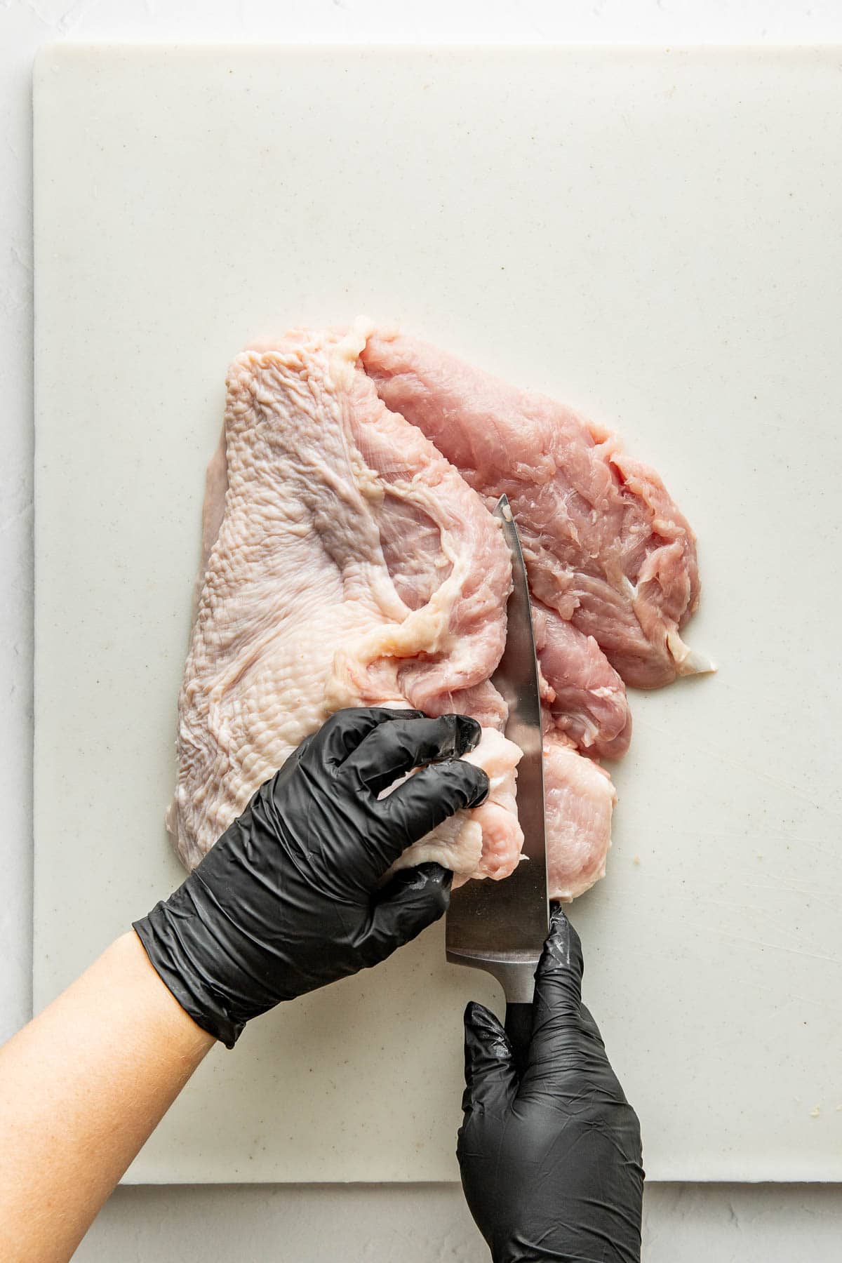 A person wearing black gloves cuts raw turkey breast on a white cutting board with a large knife.