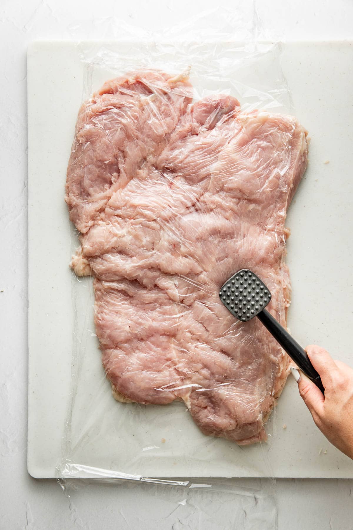 A hand uses a meat mallet to pound a large piece of raw meat covered with plastic wrap on a white cutting board.