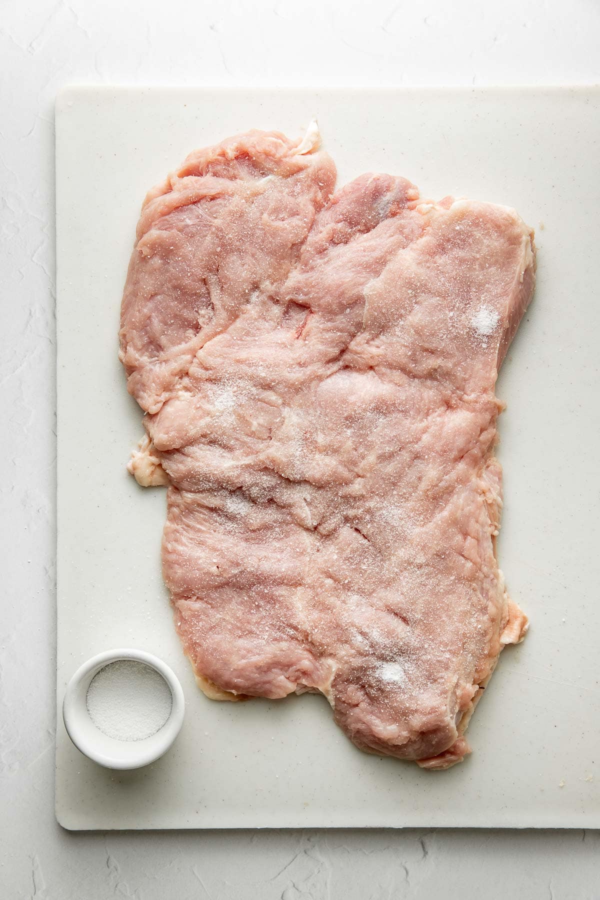 Raw, flattened chicken breast sprinkled with salt on a white cutting board, with a small bowl of salt placed beside it.