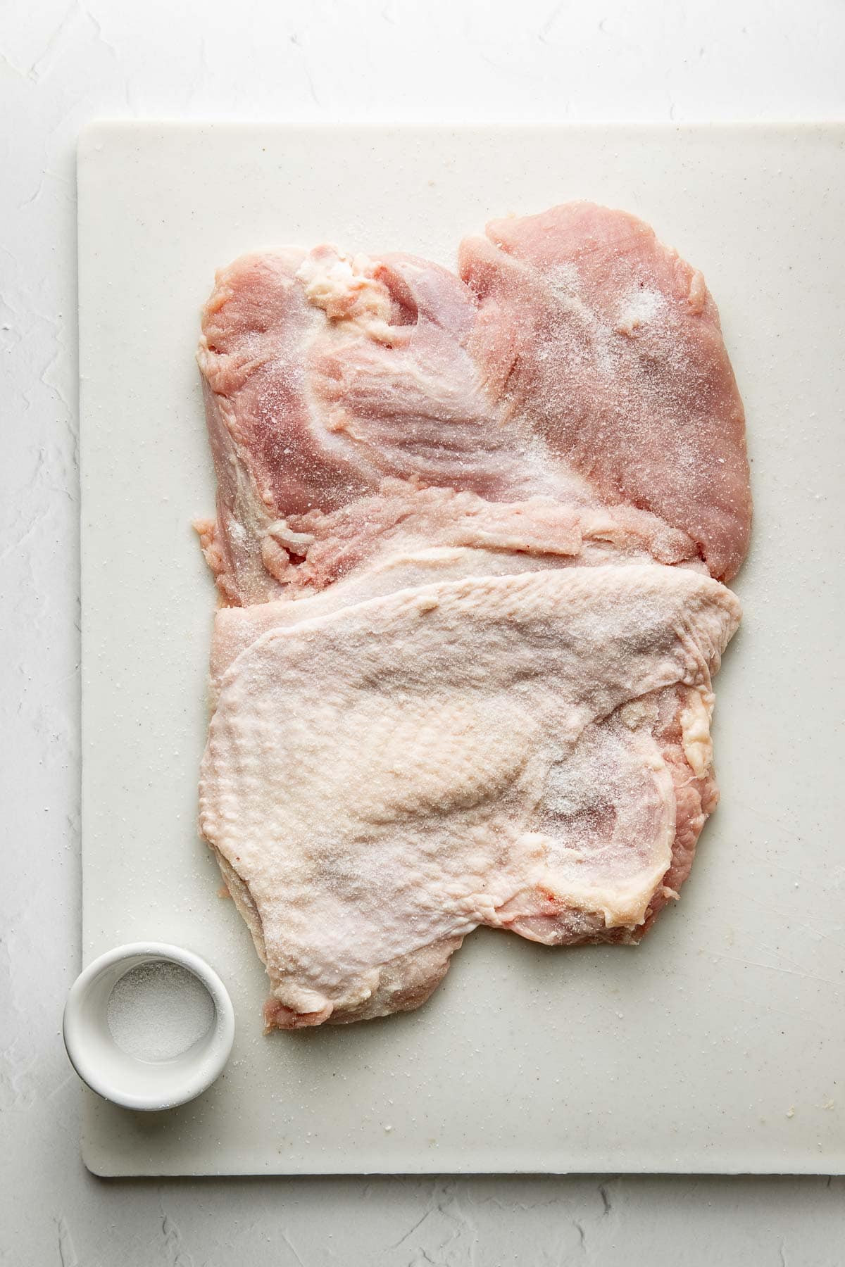 Raw turkey with skin on a white cutting board, lightly sprinkled with salt. A small white bowl filled with salt is placed beside the meat. The background is light and minimalistic.
