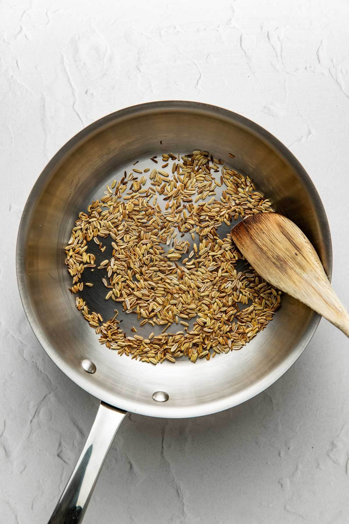 A stainless steel pan with toasted cumin seeds and a wooden spatula on a light textured surface.