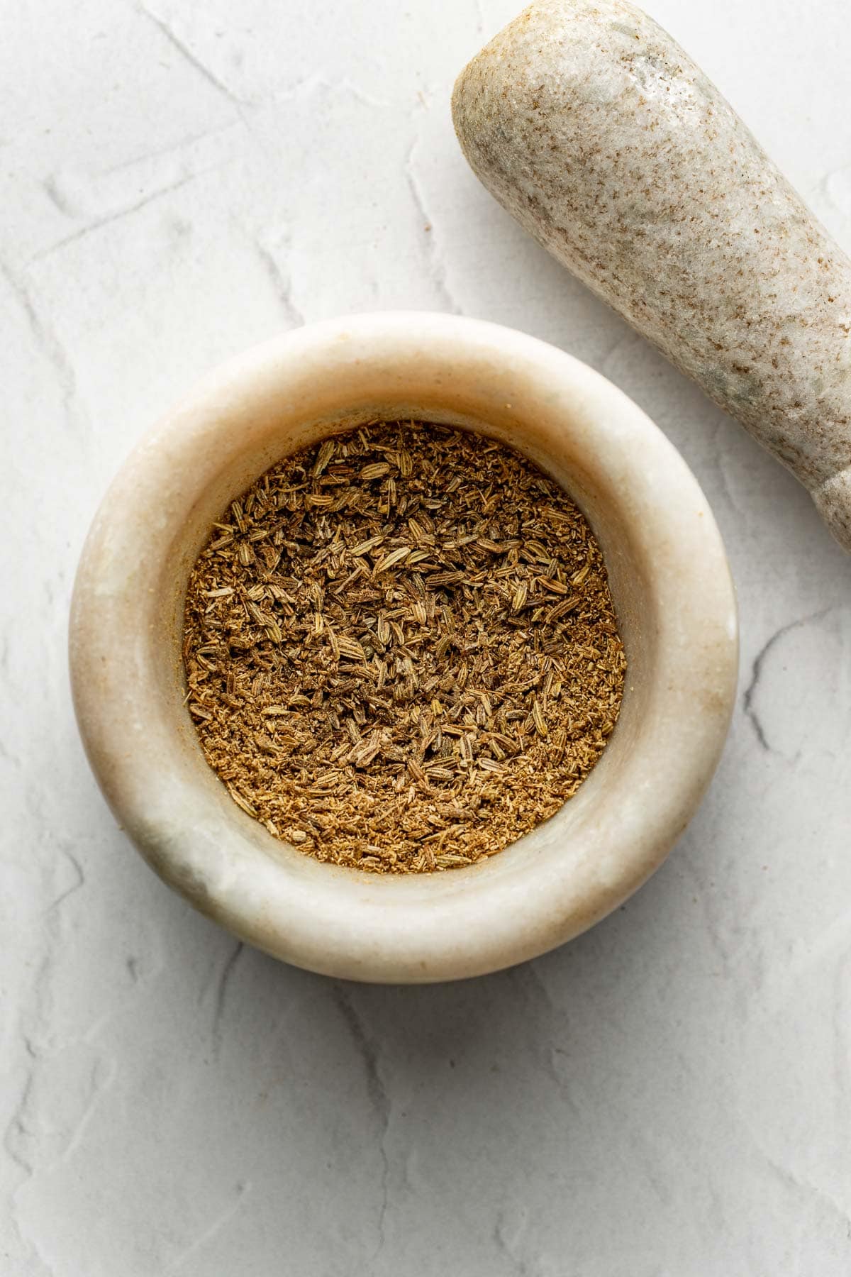 A marble mortar filled with ground brown spices sits on a light stone surface, with a matching pestle resting nearby.