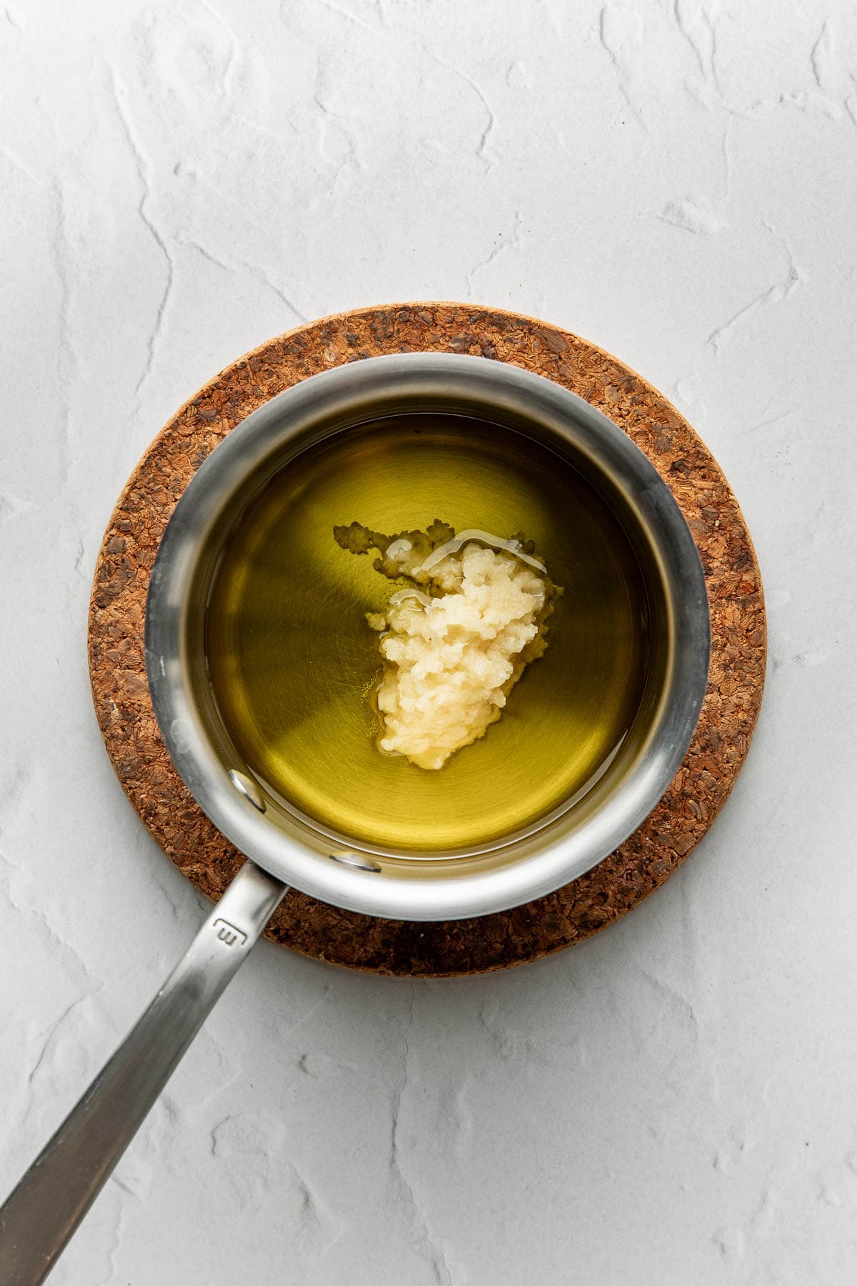 A saucepan with oil and a dollop of minced garlic sits on a cork trivet atop a light gray textured surface.