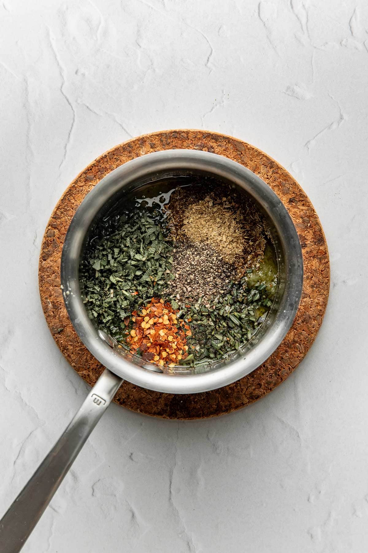 A saucepan with oil, red pepper flakes, dried herbs, and black pepper sits on a round cork trivet, placed on a light gray textured surface.