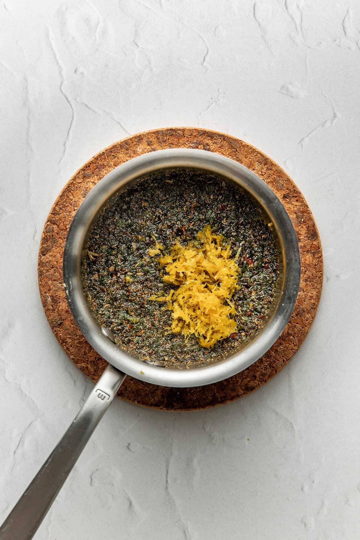 A small saucepan filled with mixed herbs and topped with fresh lemon zest sits on a round cork trivet on a light gray textured surface.
