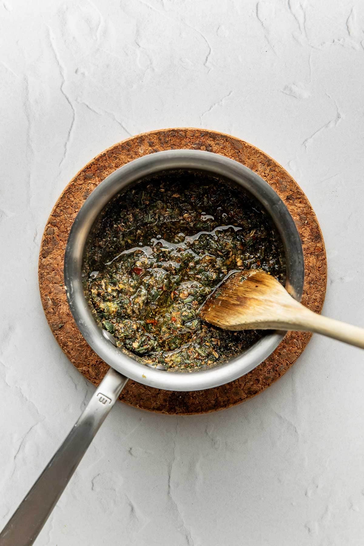 A saucepan filled with a herbed oil mixture sits on a round cork mat. A wooden spoon rests inside the pan, and the surface below is light gray with a subtle textured pattern.