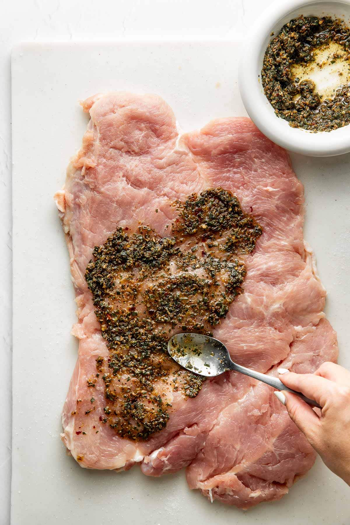 A hand uses a spoon to spread a dark herb mixture over a large, raw piece of pork on a white cutting board, with a bowl of more herb mixture nearby.