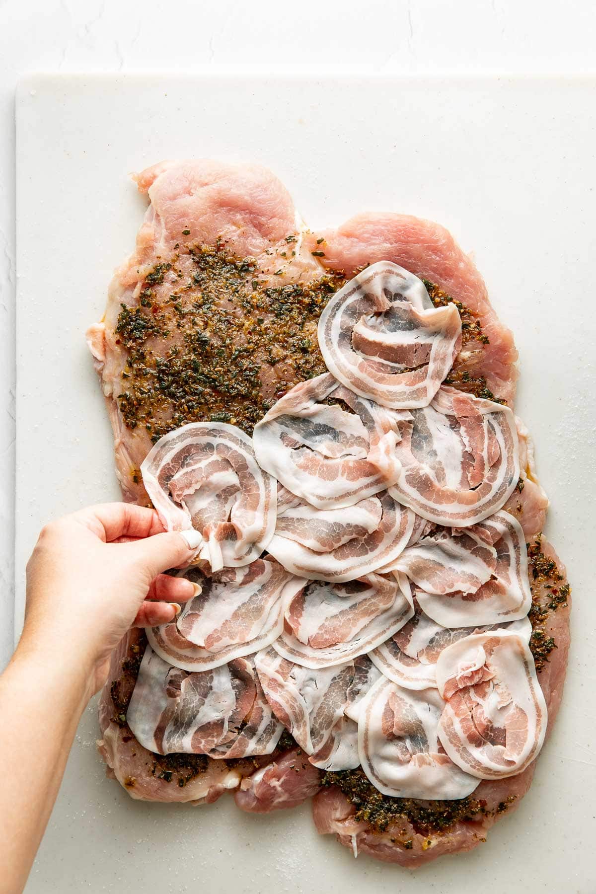 A hand places slices of pancetta over a flattened, seasoned piece of raw pork on a white cutting board.