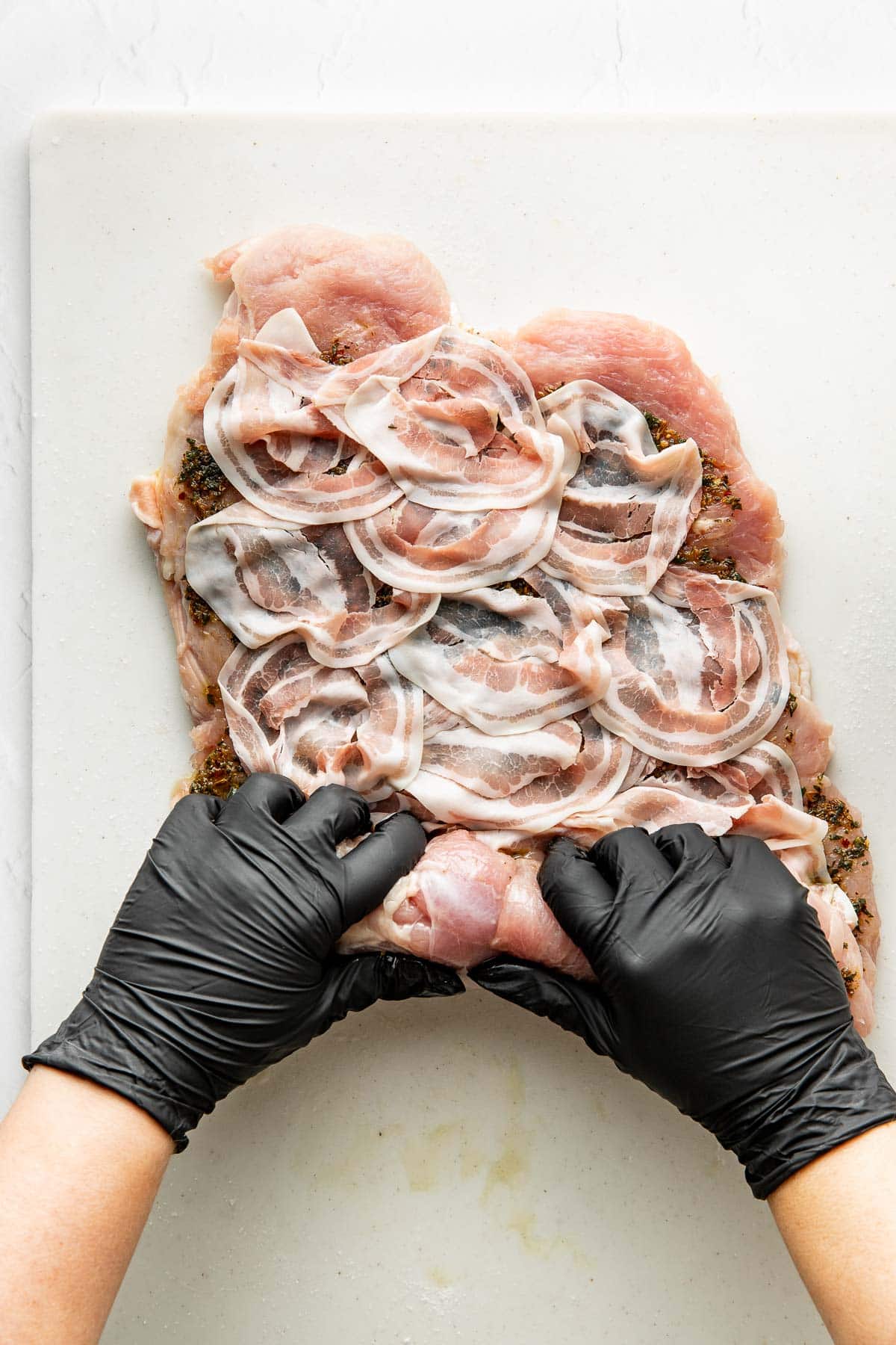 Gloved hands roll up seasoned meat layered with slices of bacon on a white cutting board, preparing it for cooking.