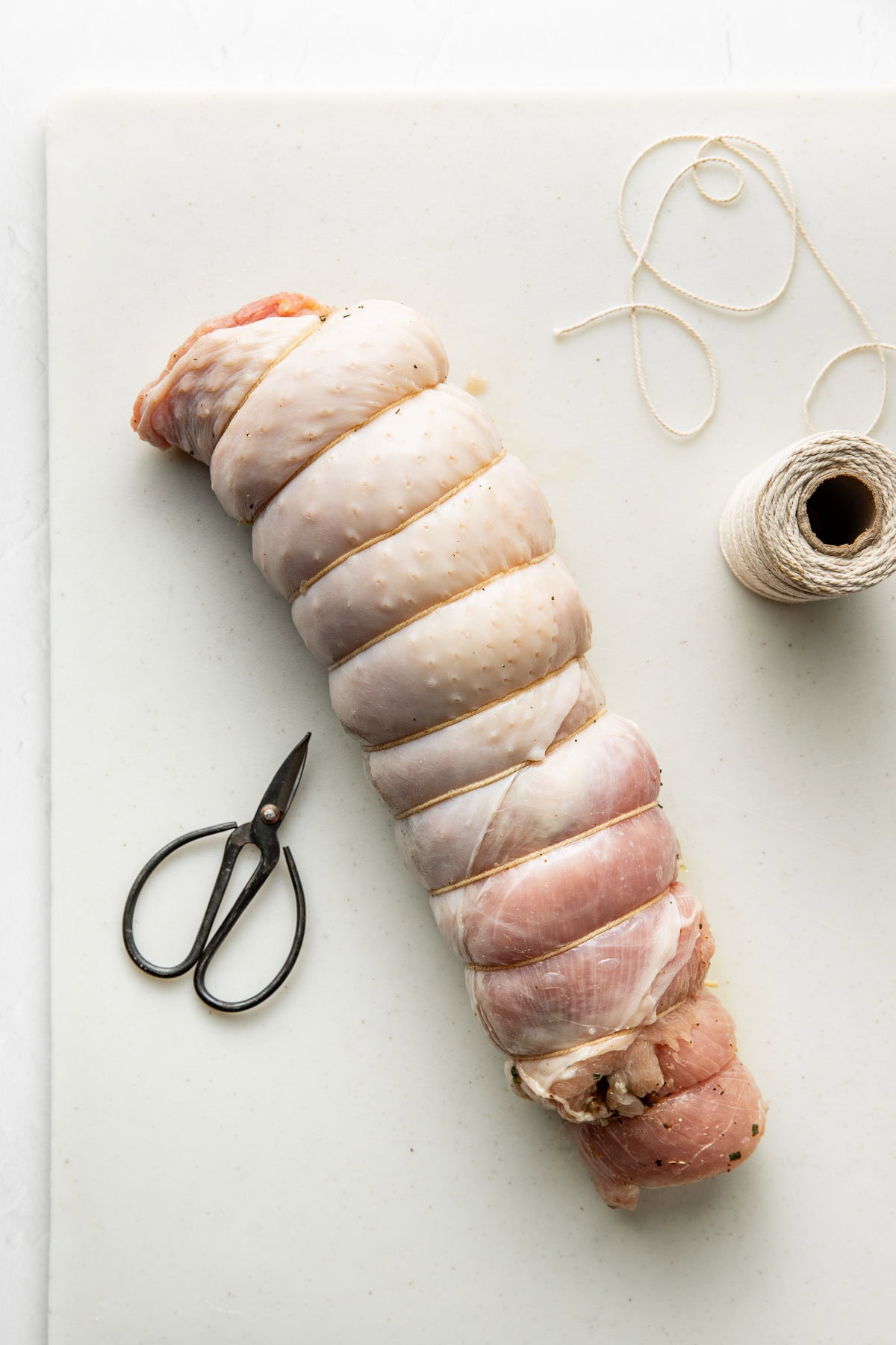 Raw, tied turkey roulade on a white cutting board next to kitchen twine and black scissors.