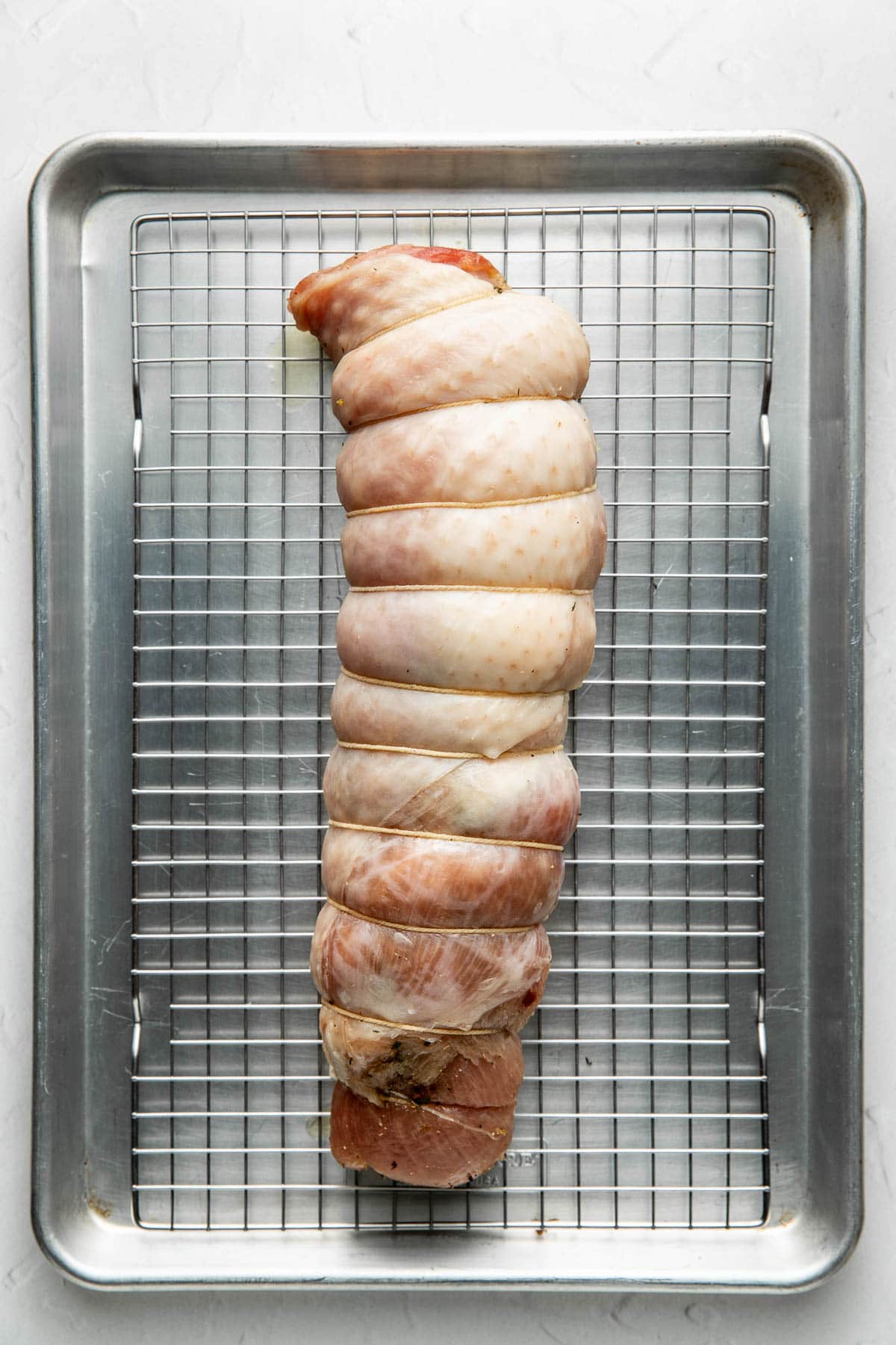 Raw, tied turkey breast roulade rests on a wire rack set over a metal baking sheet, ready for roasting, on a light textured surface.