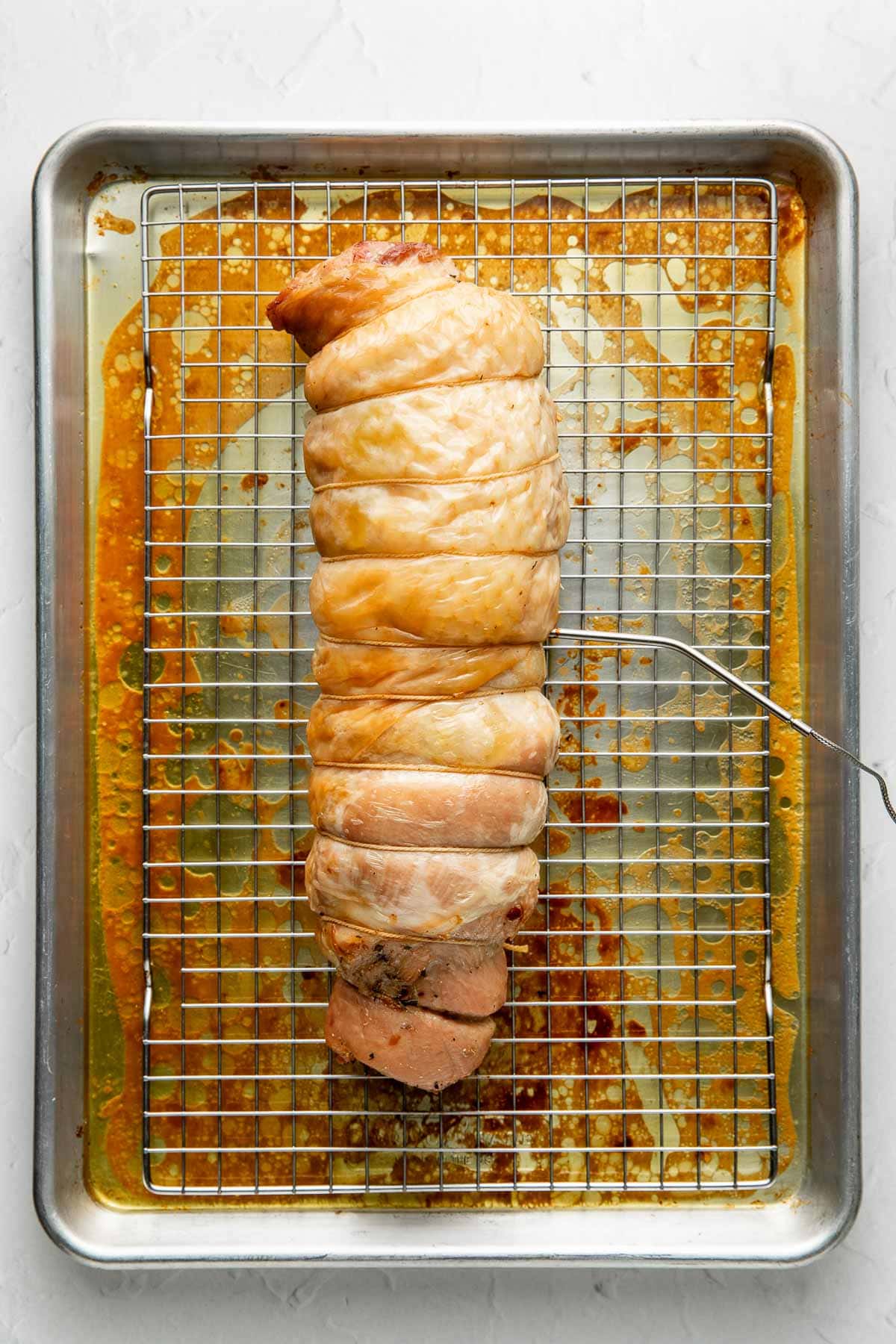 A cooked, rolled turkey breast tied with string rests on a wire rack over a baking sheet, with juices and browned bits visible beneath.