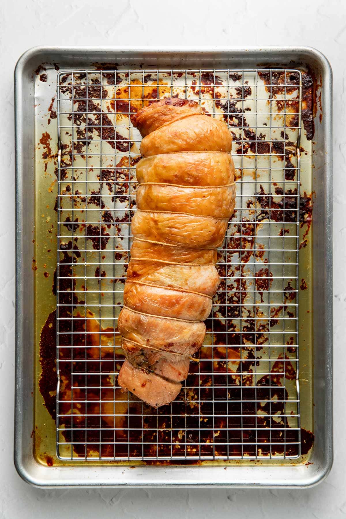 A cooked, tied turkey breast rests on a wire rack over a baking sheet with browned drippings. The turkey is golden and appears roasted, set on a light-colored background.