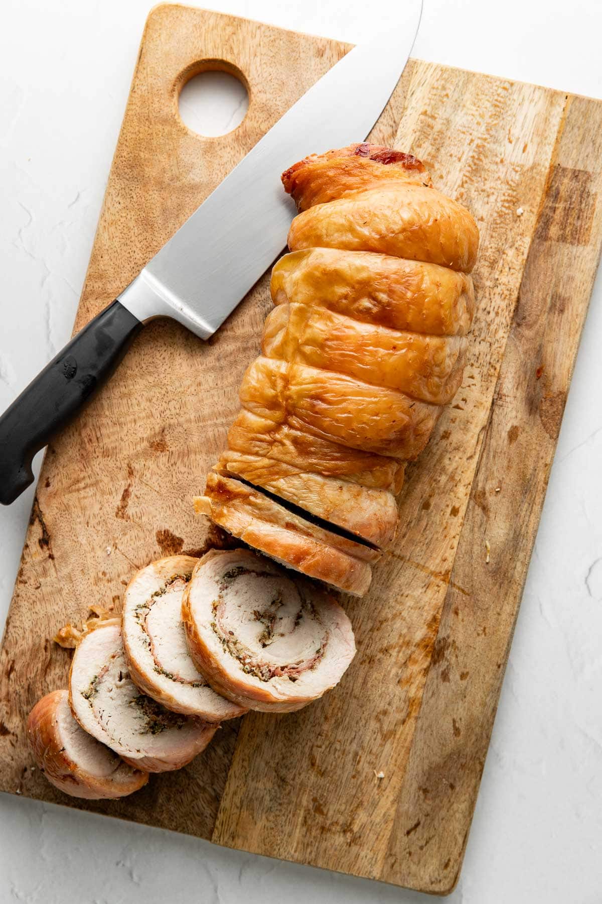 A cooked rolled roast, partially sliced, sits on a wooden cutting board next to a large kitchen knife. Three slices are cut and displayed in front of the main piece.