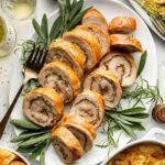 Sliced stuffed turkey roulade arranged on a white platter with fresh herbs, surrounded by side dishes like gratin potatoes, salad, dinner rolls, and glassware on a festive table.