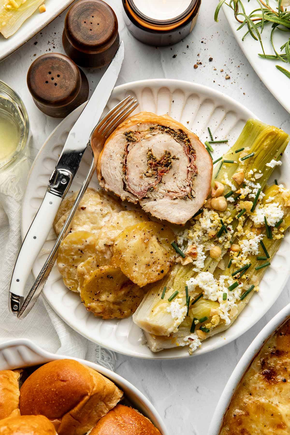 A plate with stuffed rolled meat, scalloped potatoes, leeks with cheese and herbs, a knife, and a fork. Bread rolls, salt and pepper shakers, and a glass of white wine are nearby on a white table.