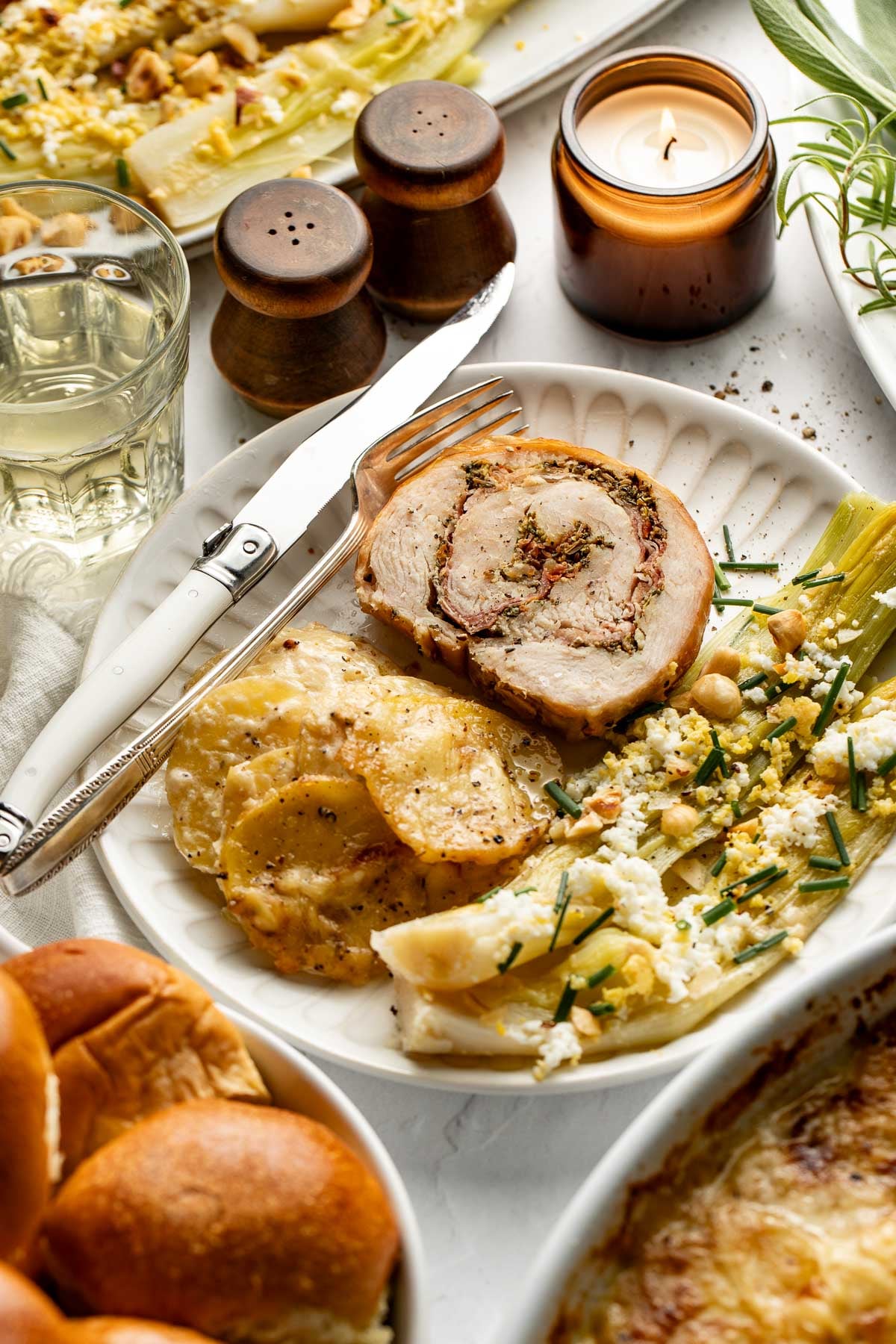 A plate with rolled stuffed meat, scalloped potatoes, and a salad sits next to bread, a glass of white wine, salt and pepper shakers, and a lit candle on a white table.