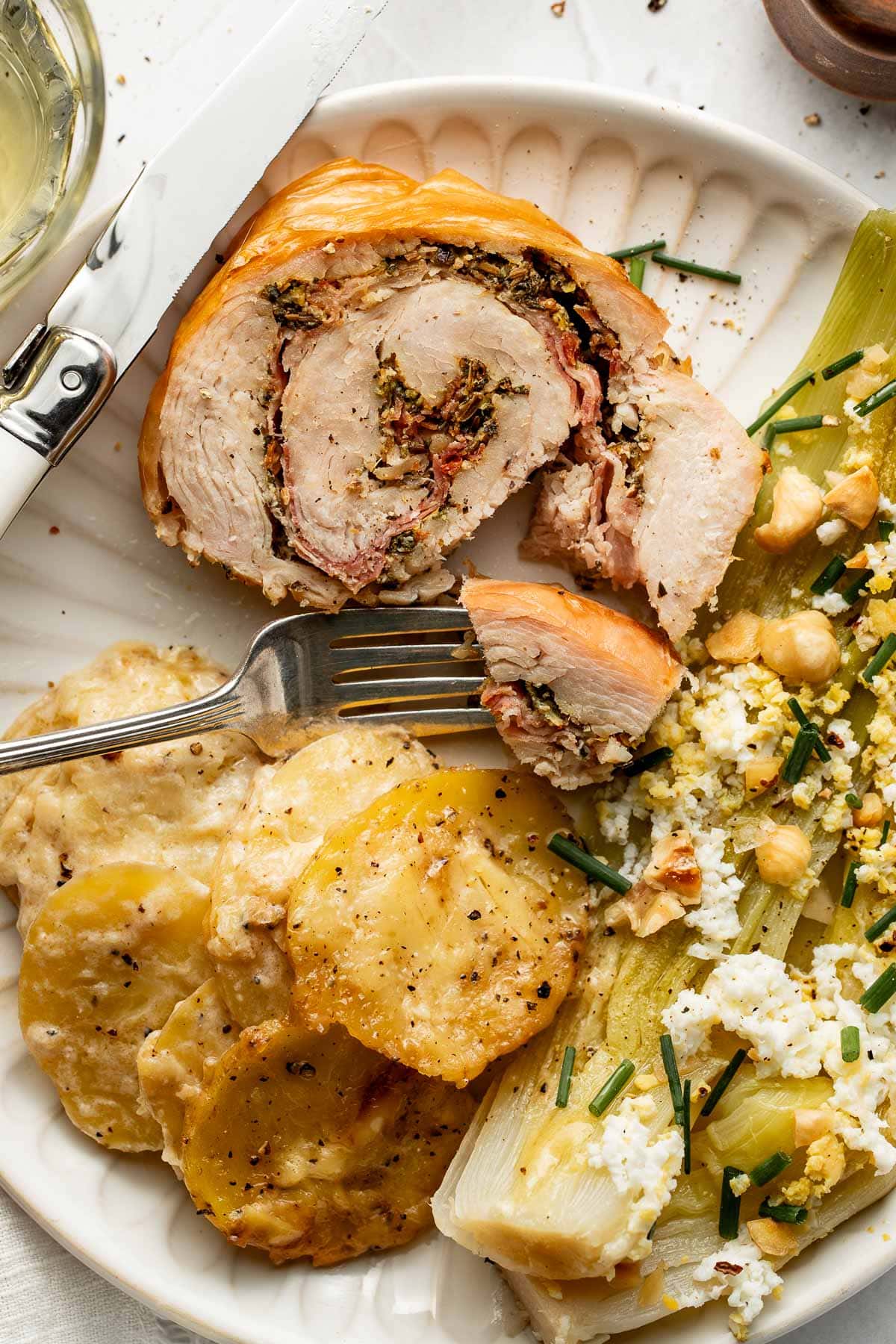 A plate with stuffed rolled chicken, sliced potatoes in a creamy sauce, and a side of vegetables topped with crumbled cheese and chives. A fork and knife rest on the plate.