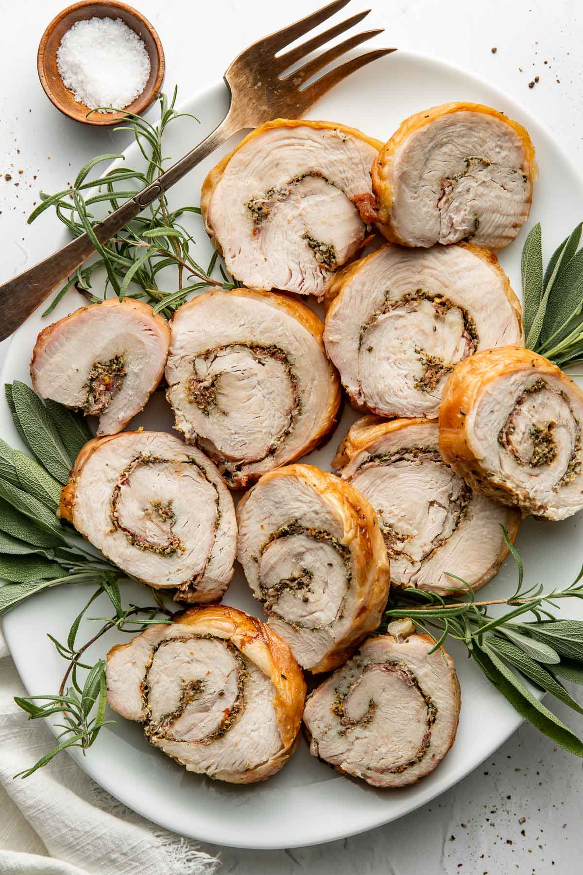 Sliced stuffed turkey roulade arranged on a white plate, garnished with fresh rosemary and sage, with a gold fork and a small bowl of salt nearby.